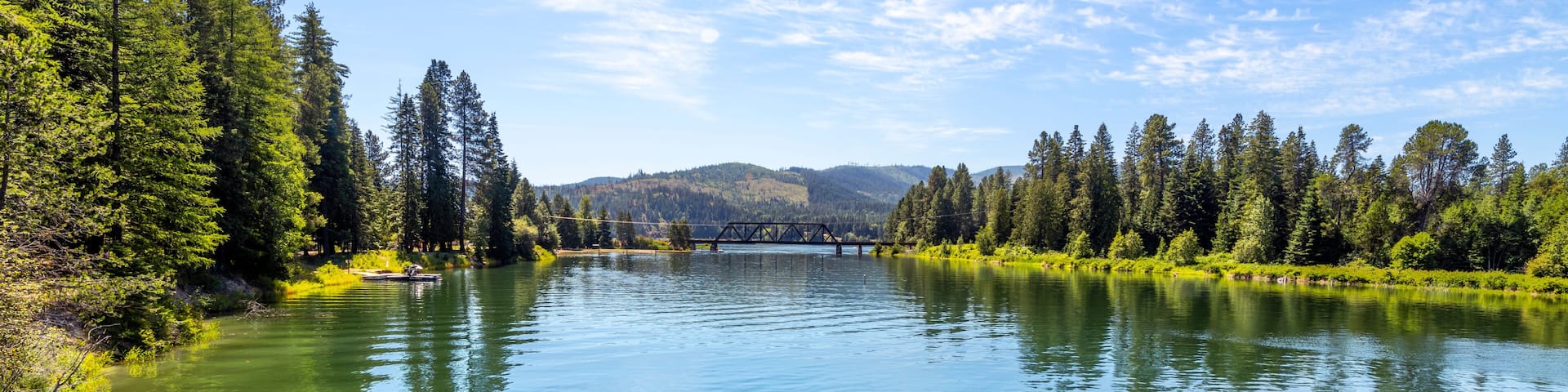 A calm river with a bridge, surrounded by dense forests and hills in the background.
