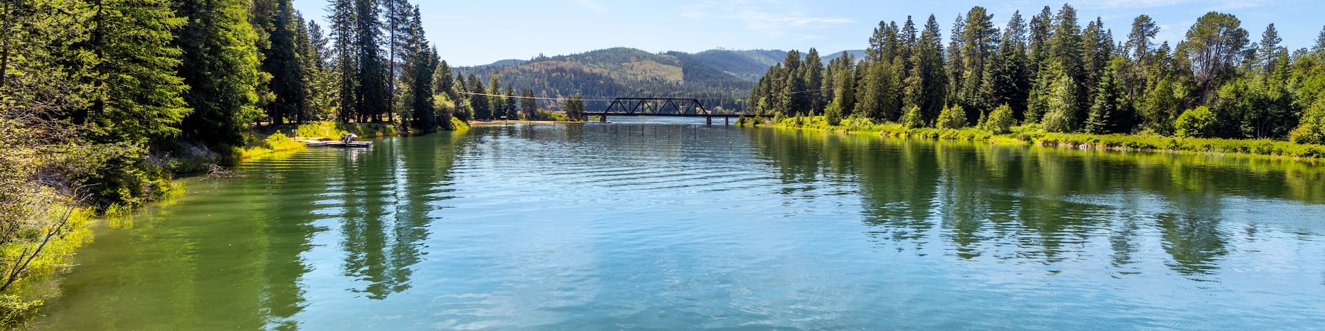 A calm river with a bridge, surrounded by dense forests and hills in the background.