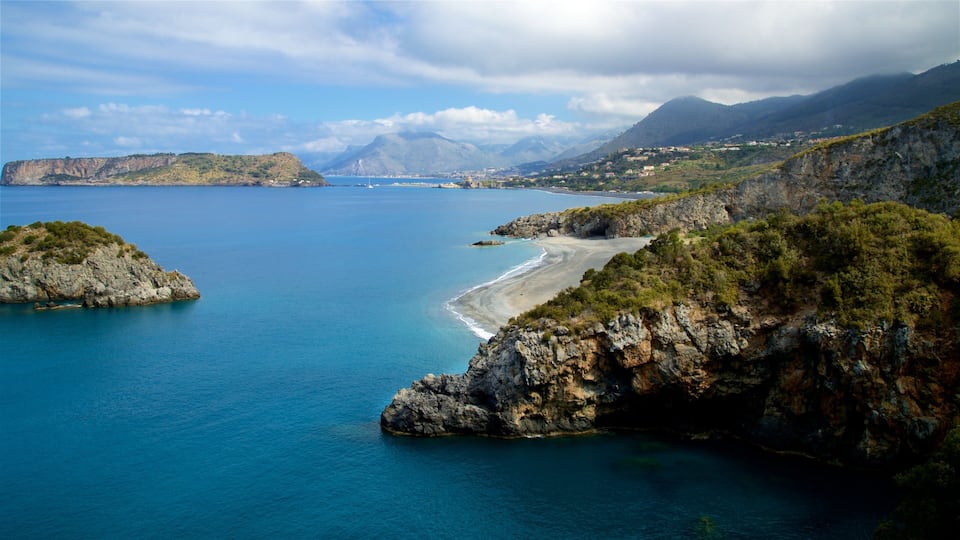 A coastal landscape with a clear blue sea, rocky cliffs, and a distant mountain range.