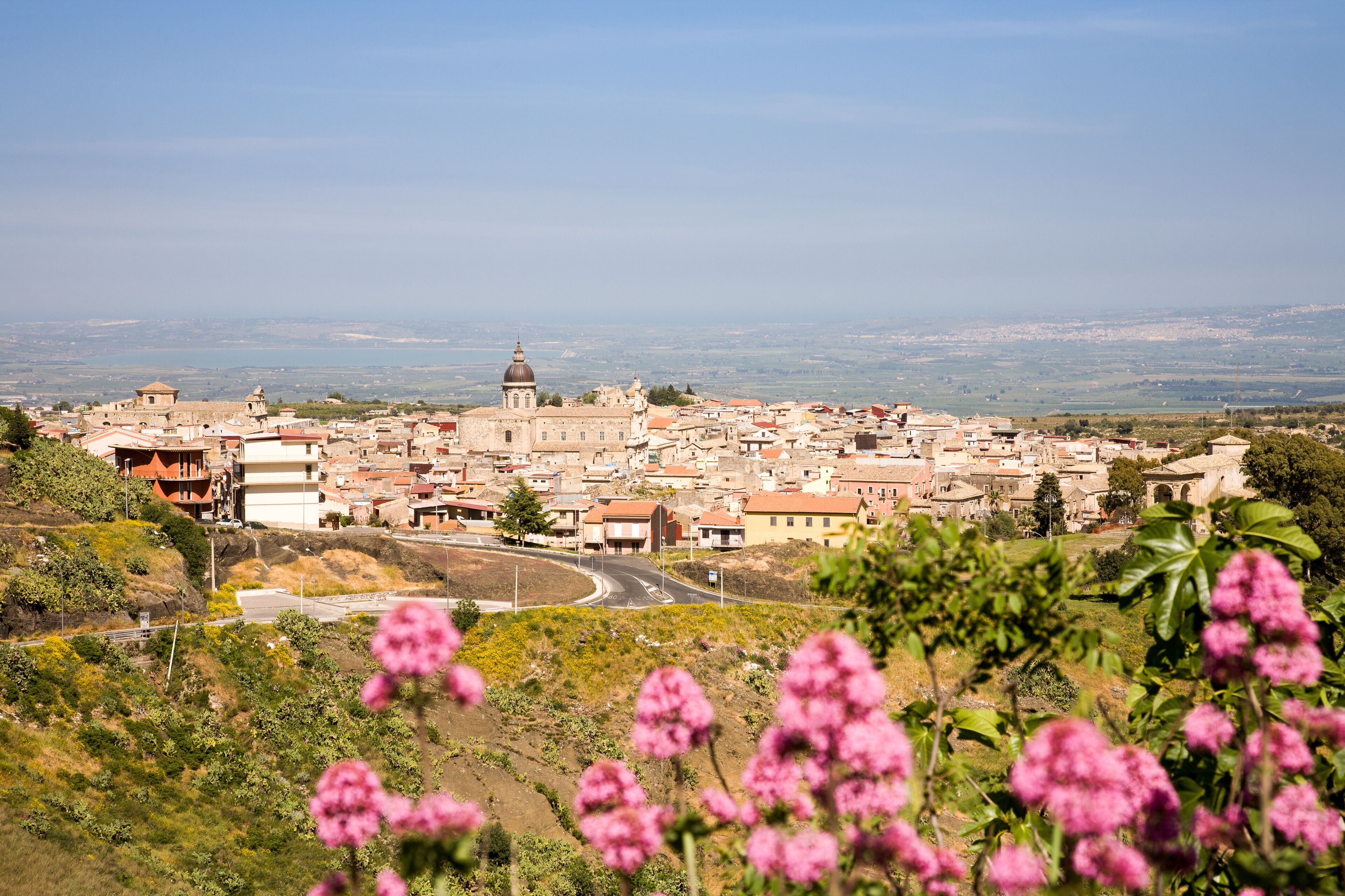 A town with a prominent church and a dome, surrounded by greenery and pink flowers.