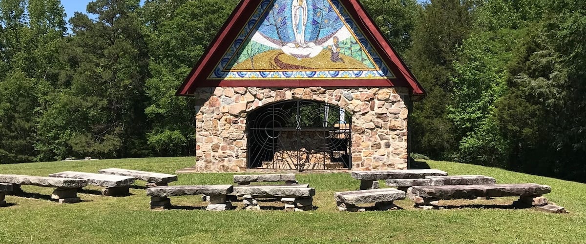 A small stone chapel with a colorful mural on the front, surrounded by a grassy field and stone benches.