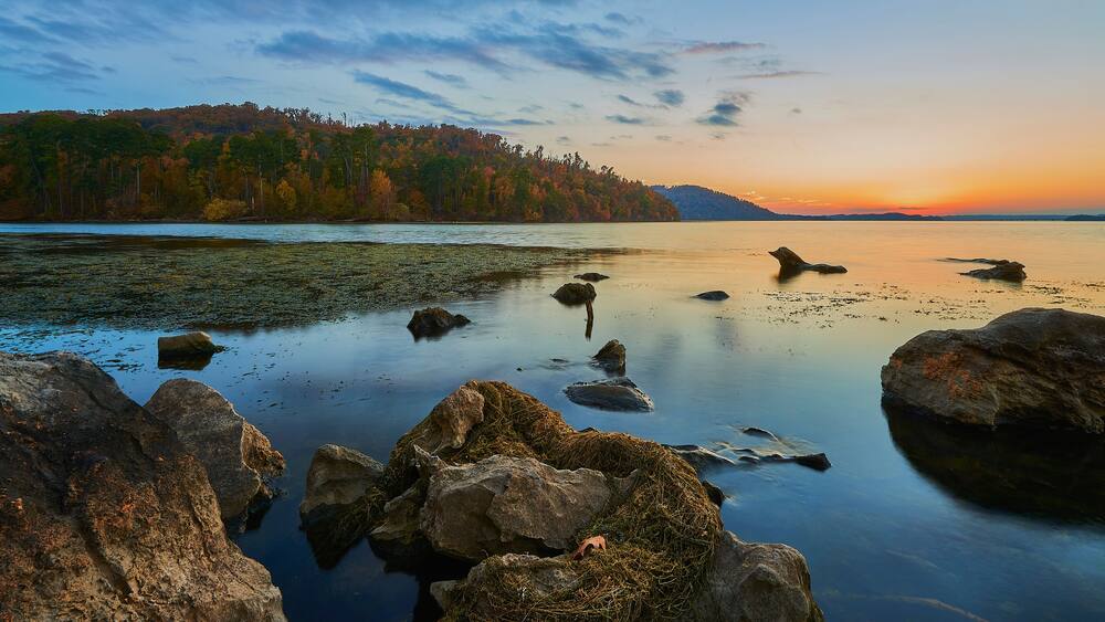 Ein ruhiger See mit Steinen im Vordergrund und einem bewaldeten Hügel im Hintergrund bei Sonnenuntergang.