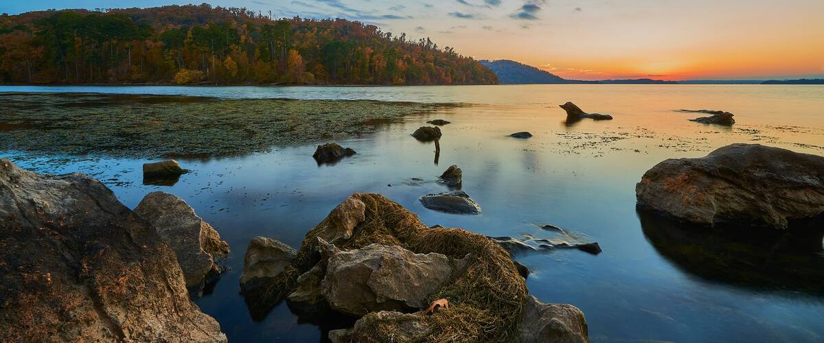A calm lake with rocks in the foreground and a forested hill in the background during sunset.