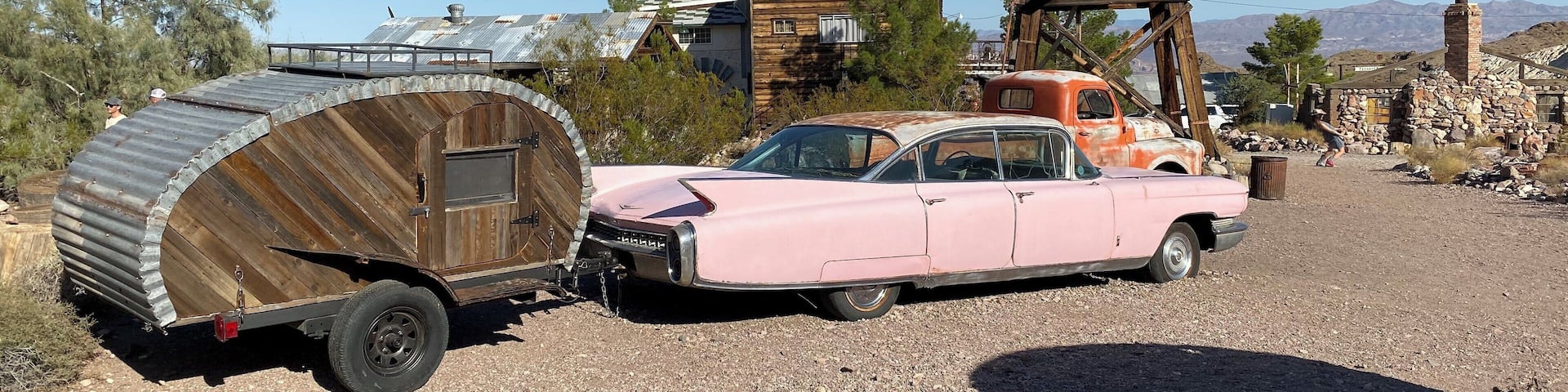 A pink vintage car with a wooden trailer in a rural setting with buildings and mountains in the background.