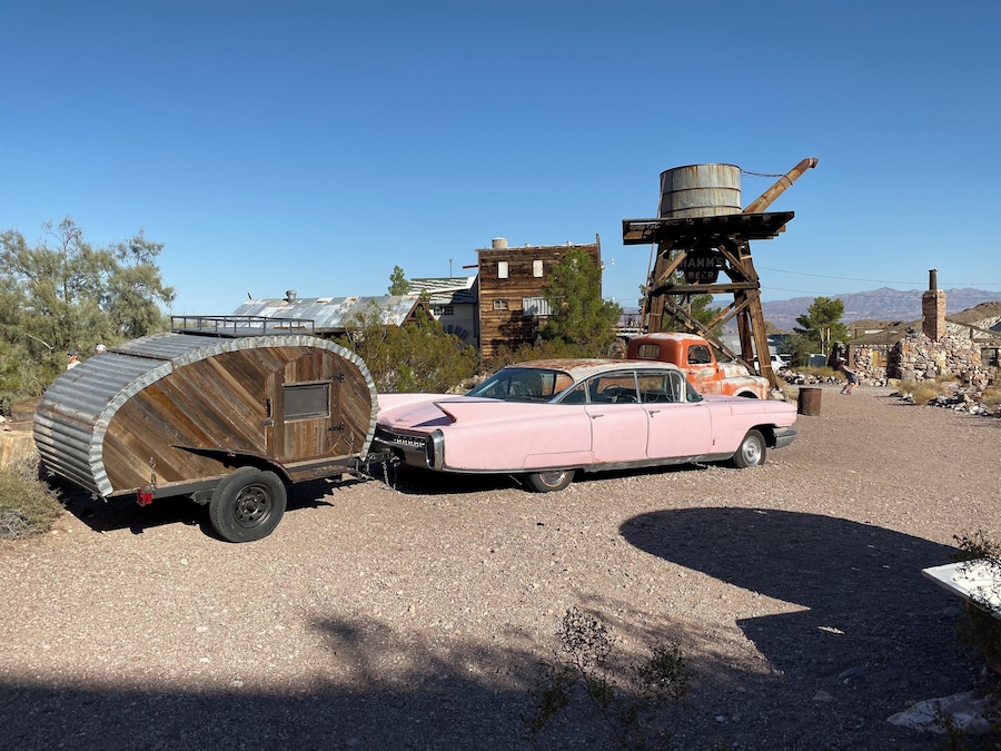 A pink vintage car with a wooden trailer in a rural setting with buildings and mountains in the background.