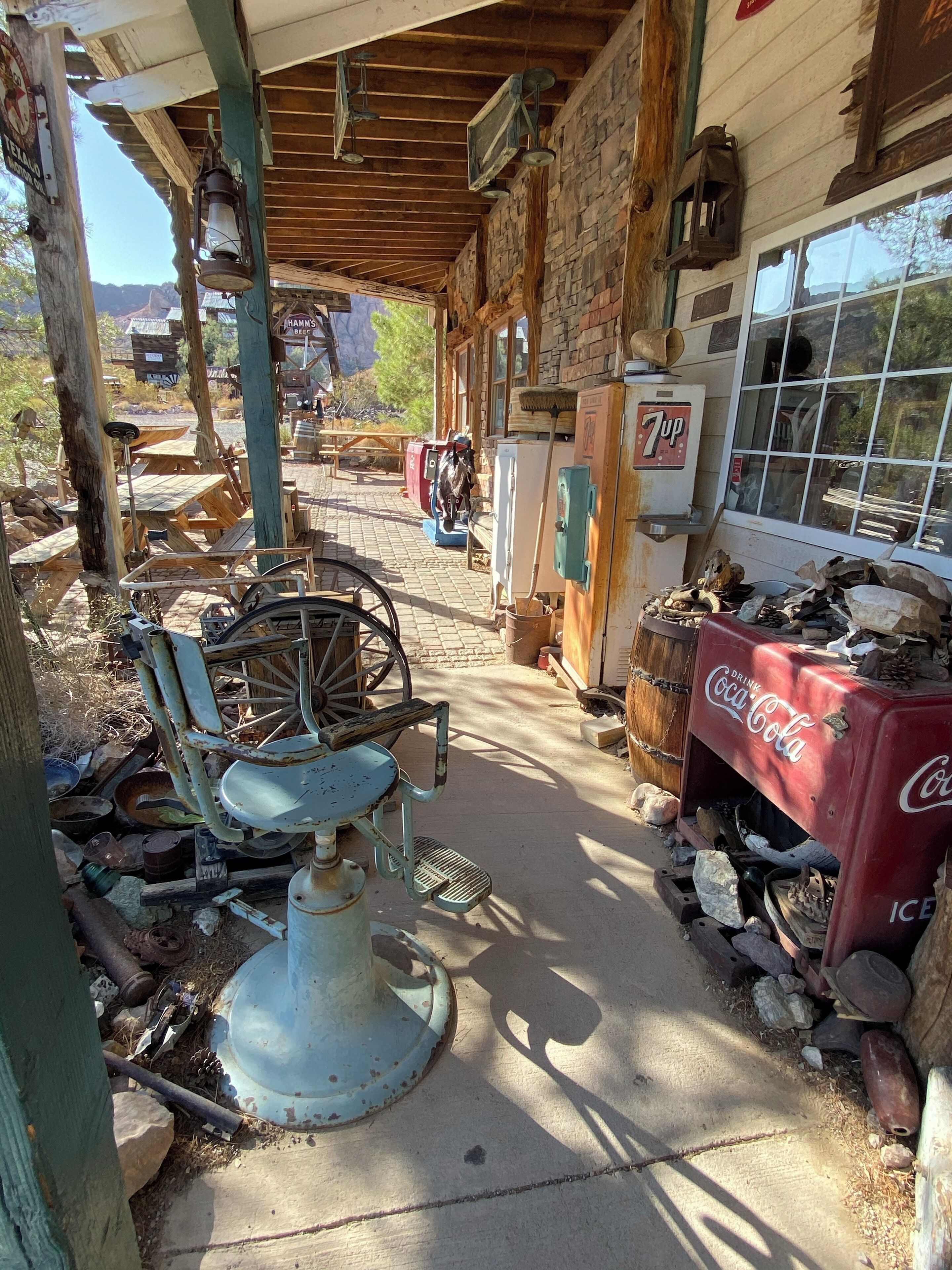 A rustic outdoor setting with a wooden structure, a vintage Coca-Cola refrigerator, and various antique items scattered around.