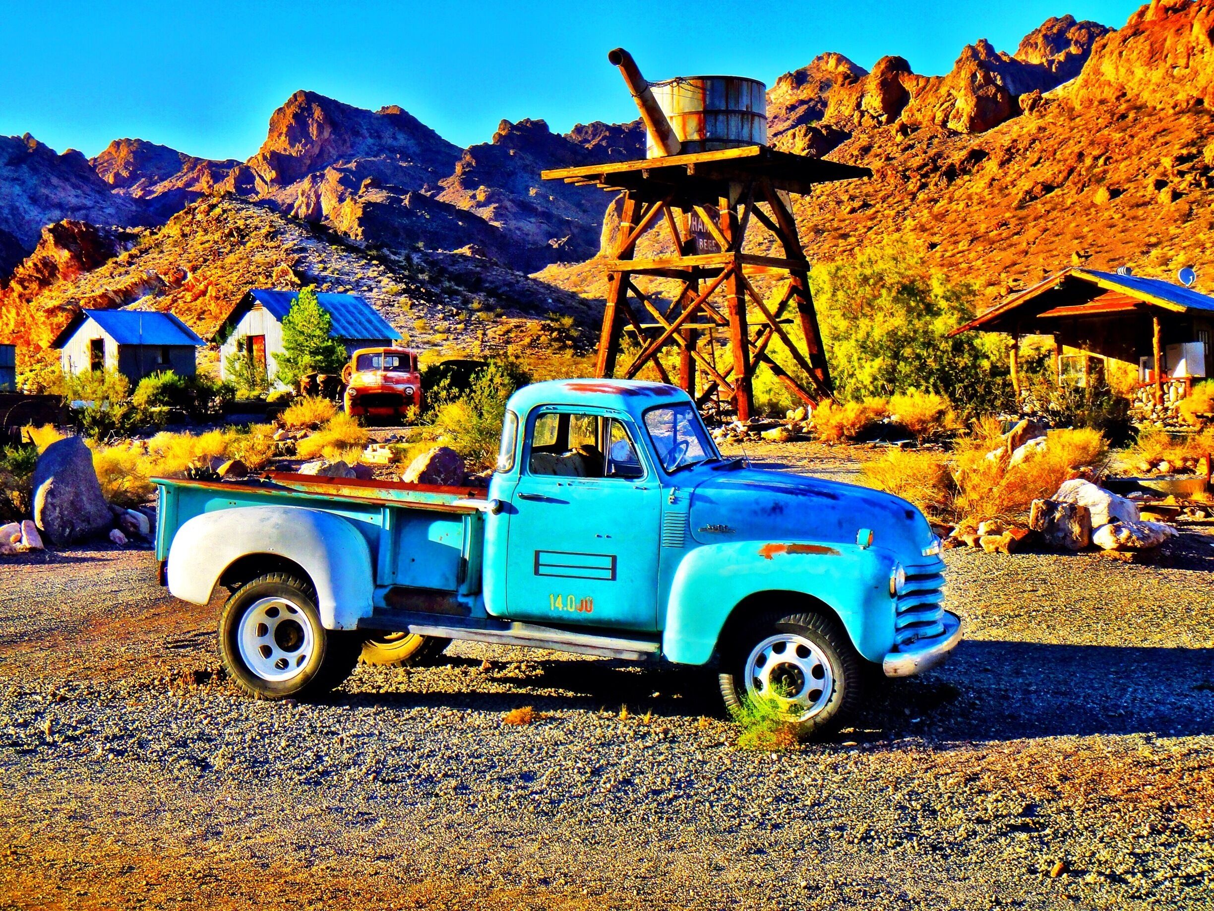A vintage turquoise truck parked in a rural area with mountains in the background.