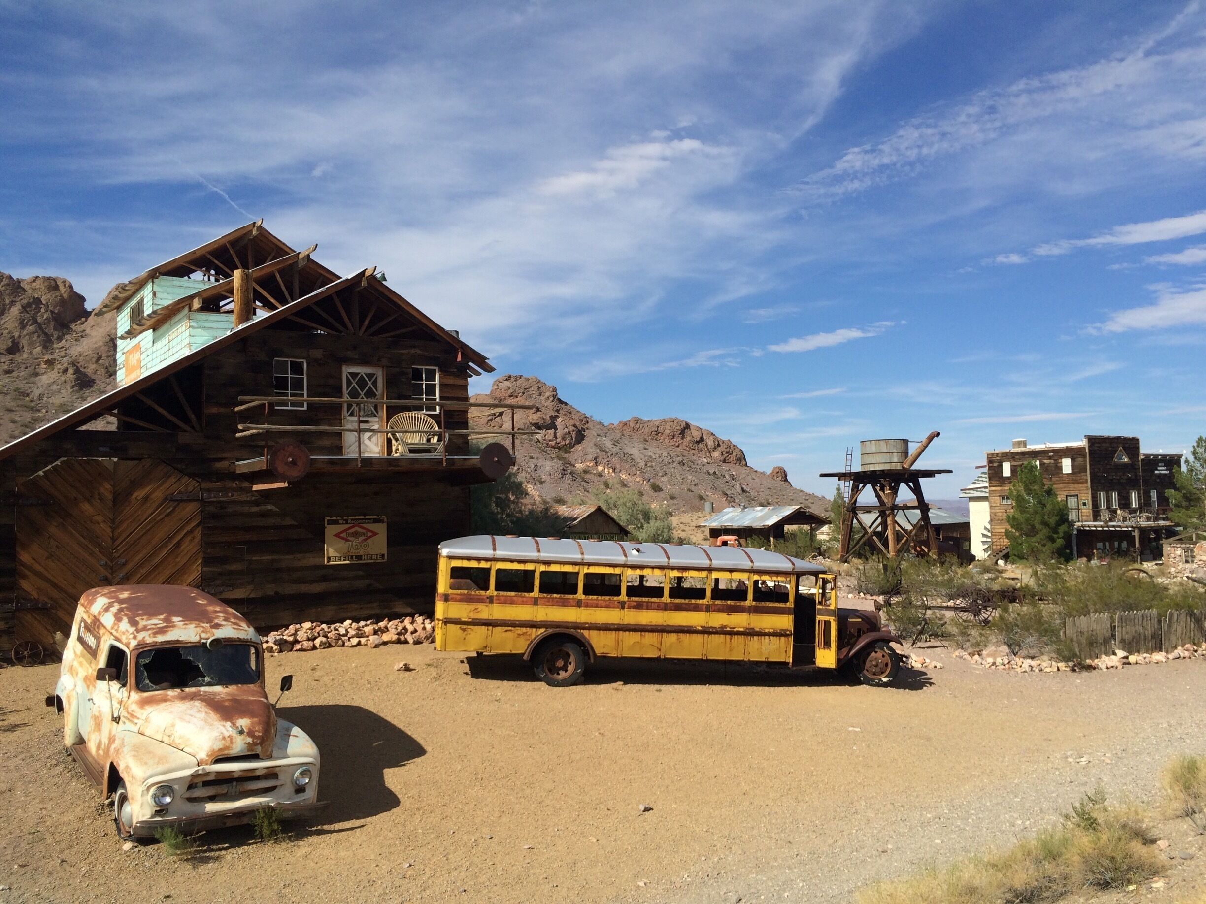 A rustic wooden building, a vintage yellow bus, and an old car in a desert setting.