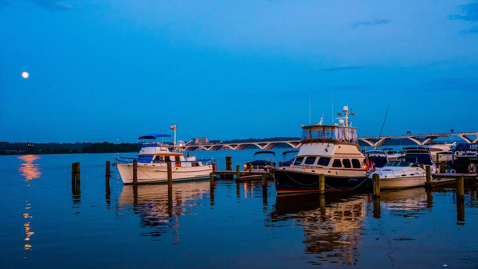A marina at dusk with several boats docked, a bridge in the background, and a calm water surface reflecting the sky.