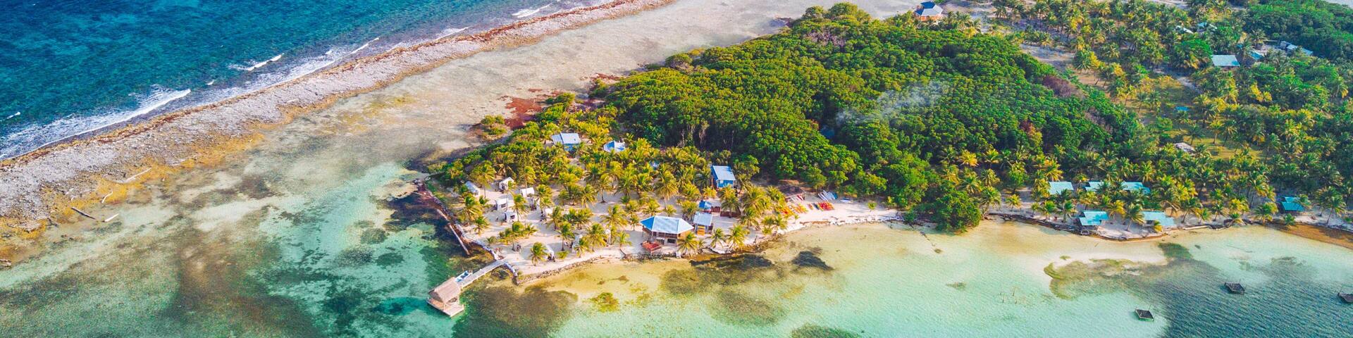 An aerial view of a tropical island with clear turquoise waters, a sandy beach, and a cluster of small buildings surrounded by dense green foliage.
