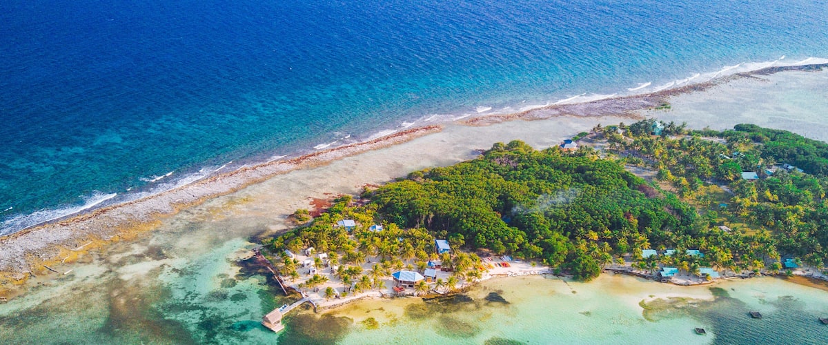 An aerial view of a tropical island with clear turquoise waters, a sandy beach, and a cluster of small buildings surrounded by dense green foliage.