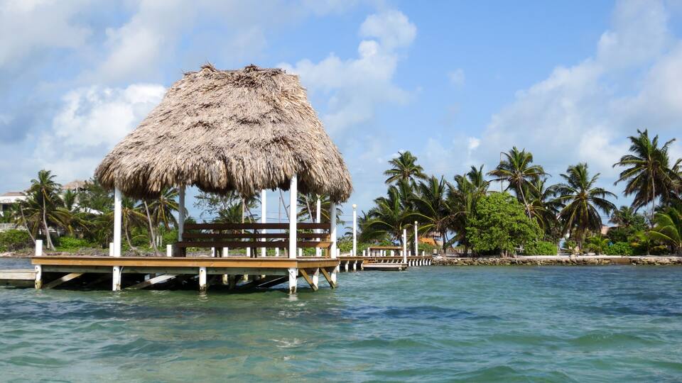 A thatched-roof hut on stilts over turquoise water, surrounded by palm trees and a clear sky.