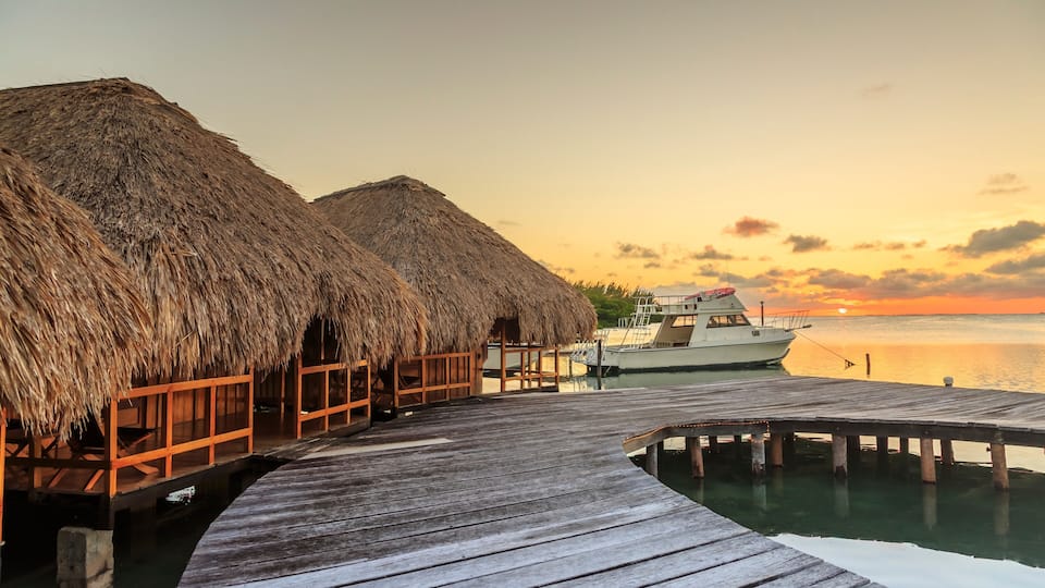A wooden pier leading to overwater bungalows with thatched roofs, a boat docked at the end, and a sunset over calm waters.