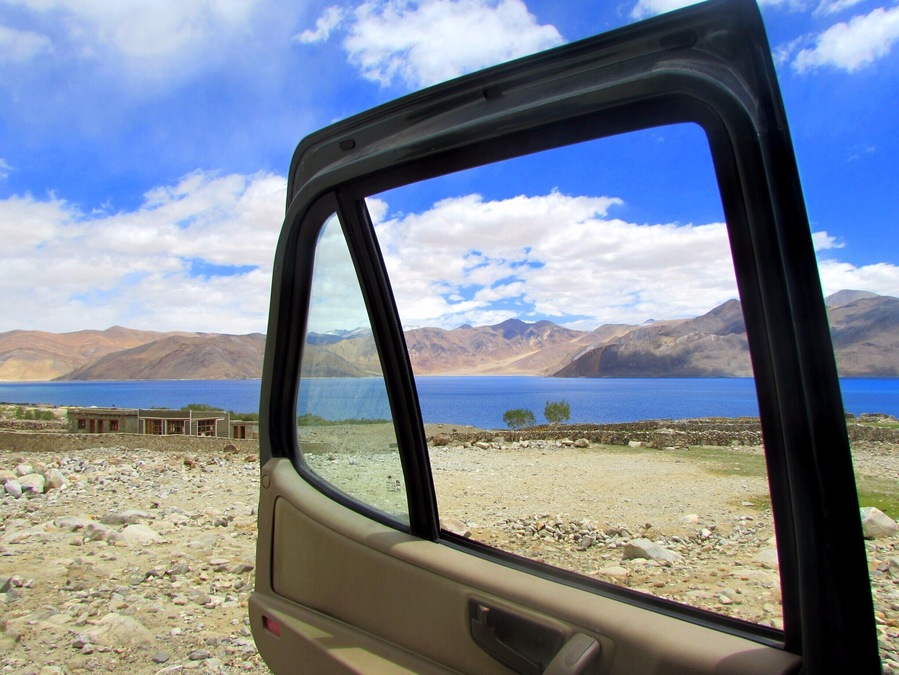Ventana lateral de un coche con vistas a un lago, montañas y un cielo despejado.
