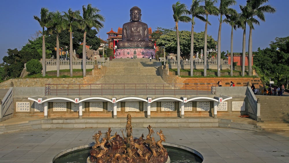 A large Buddha statue, palm trees, and a fountain with sculptures.
