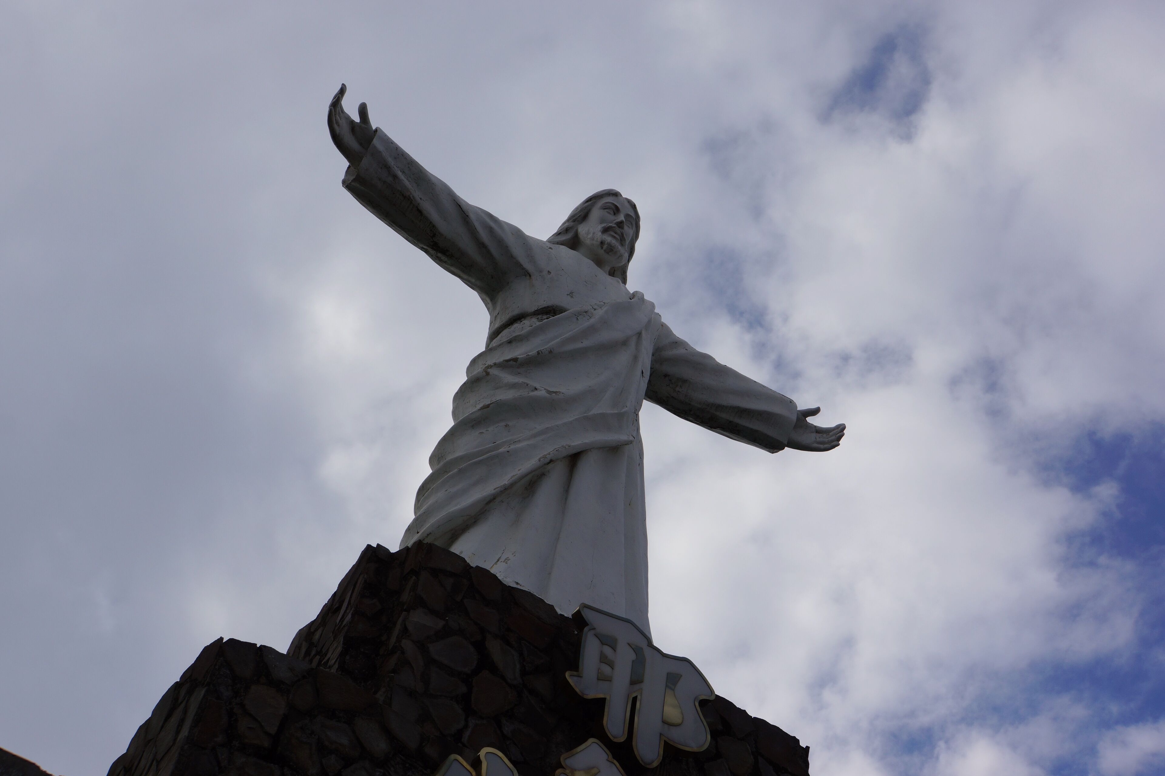 A statue with outstretched arms on a rocky pedestal against a cloudy sky.