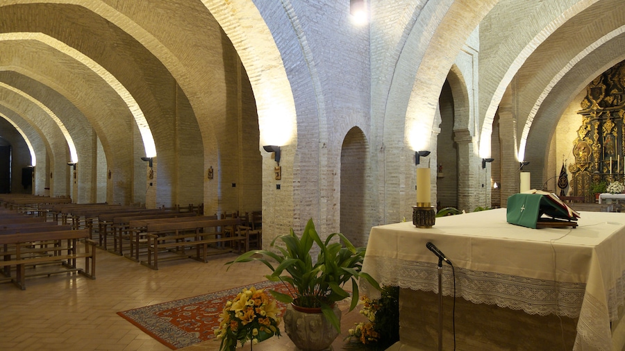 Interior de una iglesia con techos abovedados, bancales de madera, un atril y un gran crucifijo.