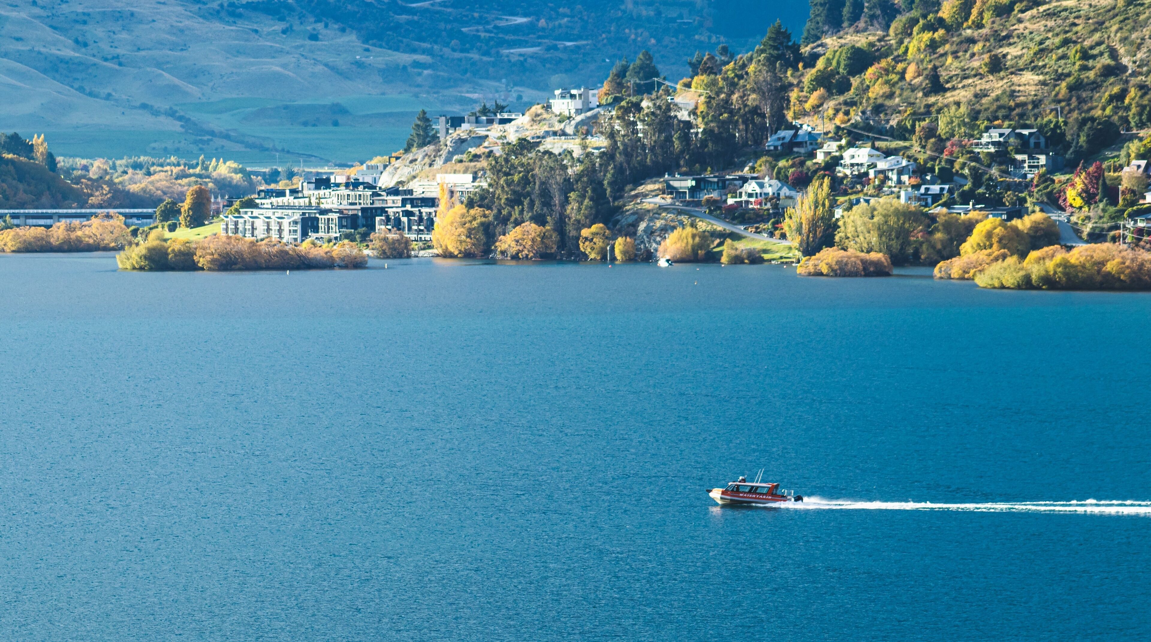A lake with a boat, houses on a hillside, and autumn trees.