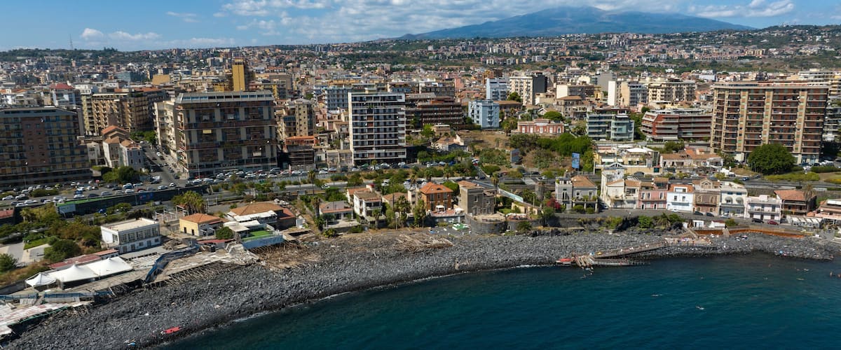 A coastal cityscape with a rocky shoreline, dense urban buildings, and a distant mountain range.