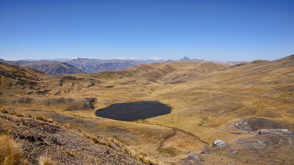 A dry lake with a small pond surrounded by barren hills and distant snow-capped mountains.