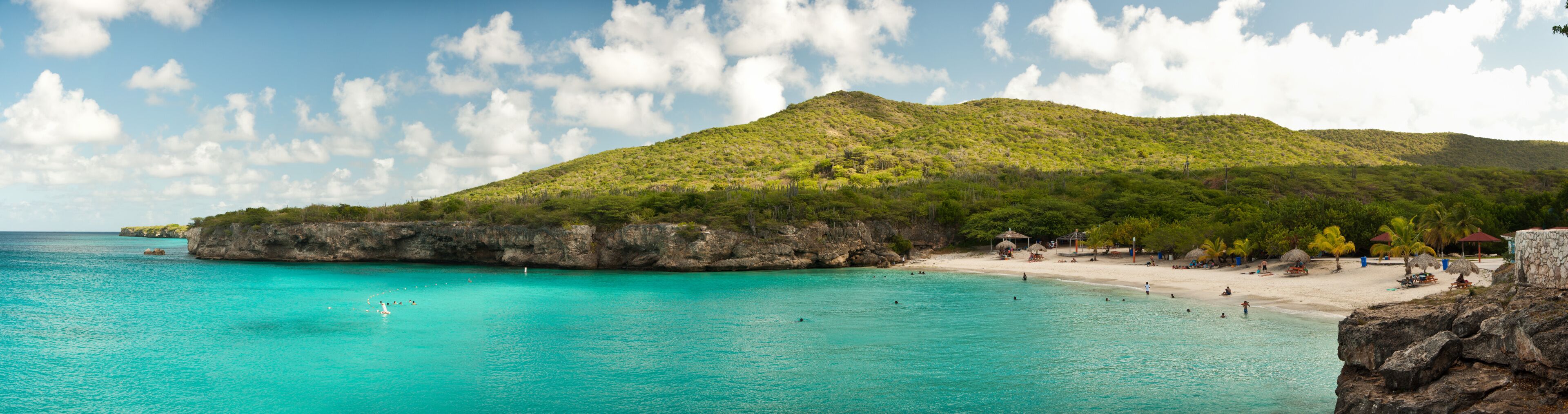 A turquoise beach with clear waters, a rocky coastline, and a lush green hill in the background.