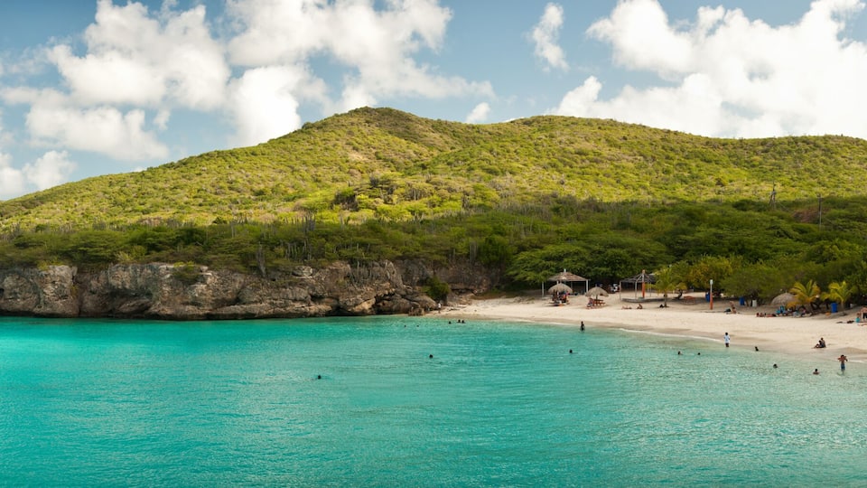 A turquoise beach with clear waters, a rocky coastline, and a lush green hill in the background.
