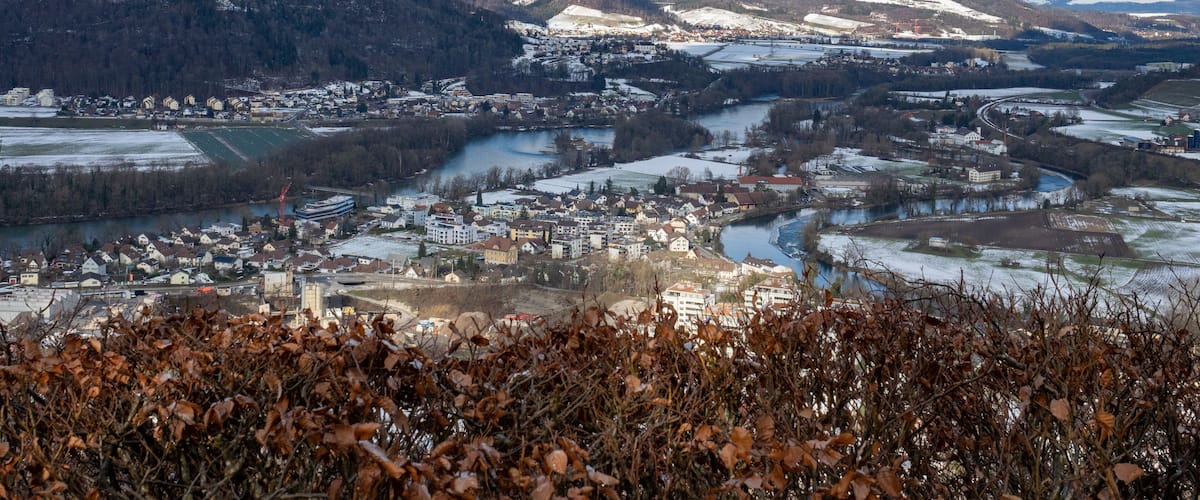 A winter landscape with a town nestled by a river, snow-covered hills, and a clear sky.