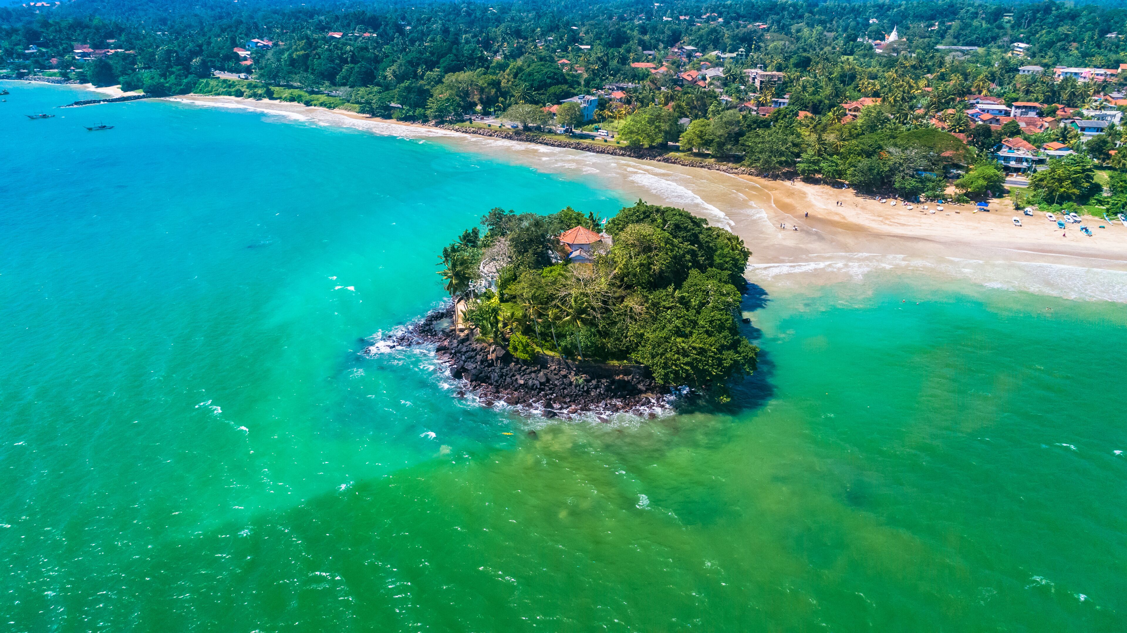 An aerial view of a tropical island surrounded by turquoise waters, with a sandy beach and dense greenery.
