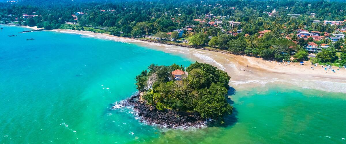 An aerial view of a tropical island surrounded by turquoise waters, with a sandy beach and dense greenery.