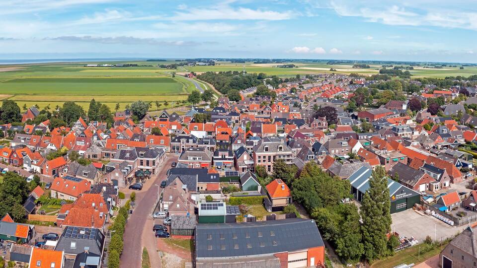 An aerial view of a town with red-roofed houses, a church with a tall steeple, and expansive green fields in the background.