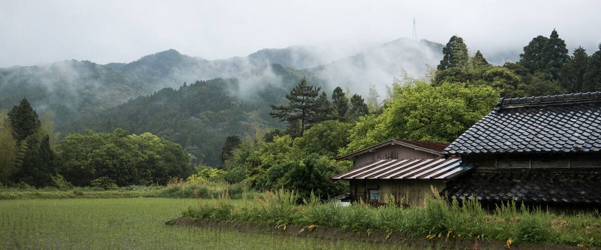 A traditional Japanese house by a serene lake, surrounded by lush greenery and mist-covered mountains.