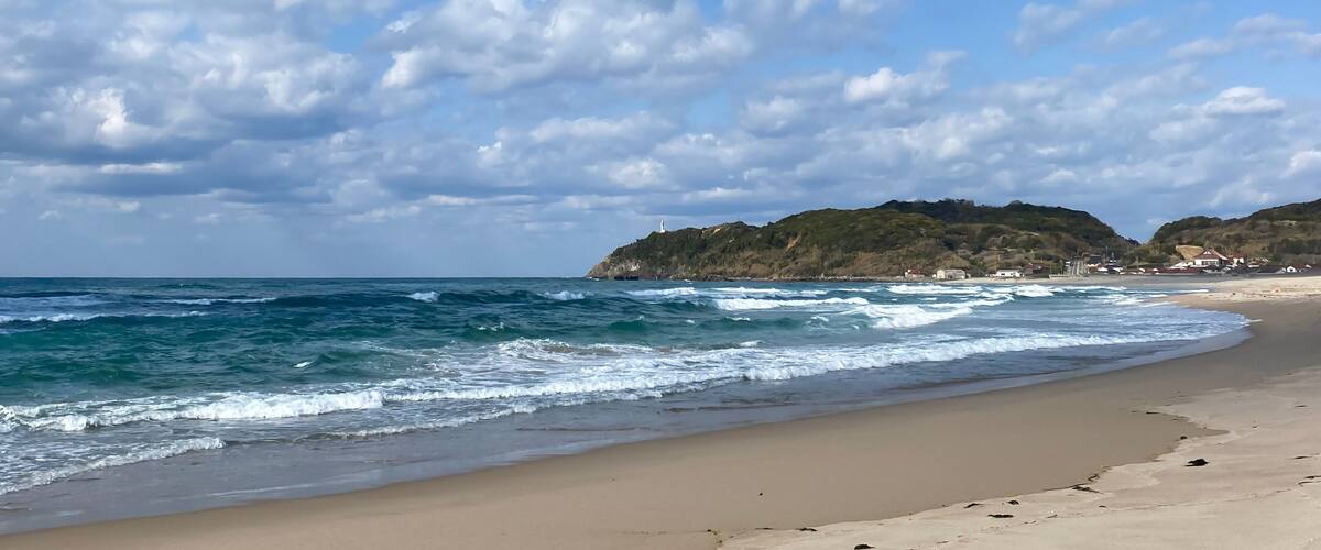 A beach with waves, a clear sky, and a hill with buildings.