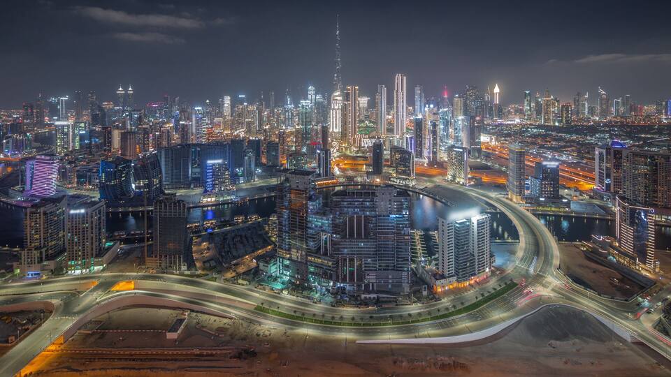A nighttime cityscape with illuminated skyscrapers, a prominent tower, and a complex network of roads.