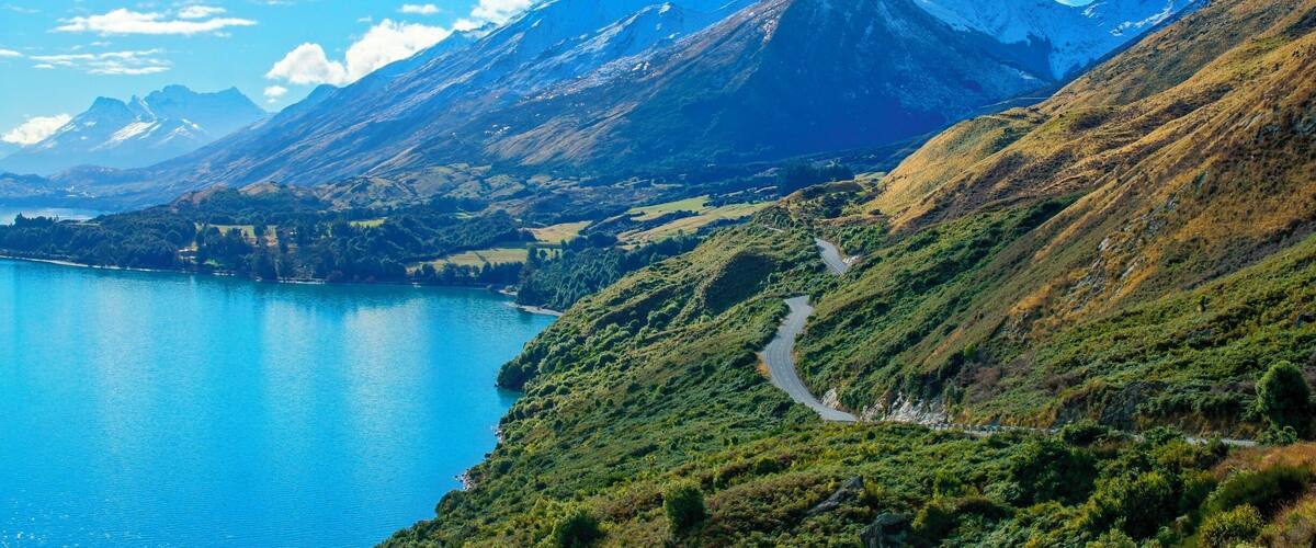 A mountainous landscape with a winding road, a lake, and snow-capped peaks under a clear blue sky.