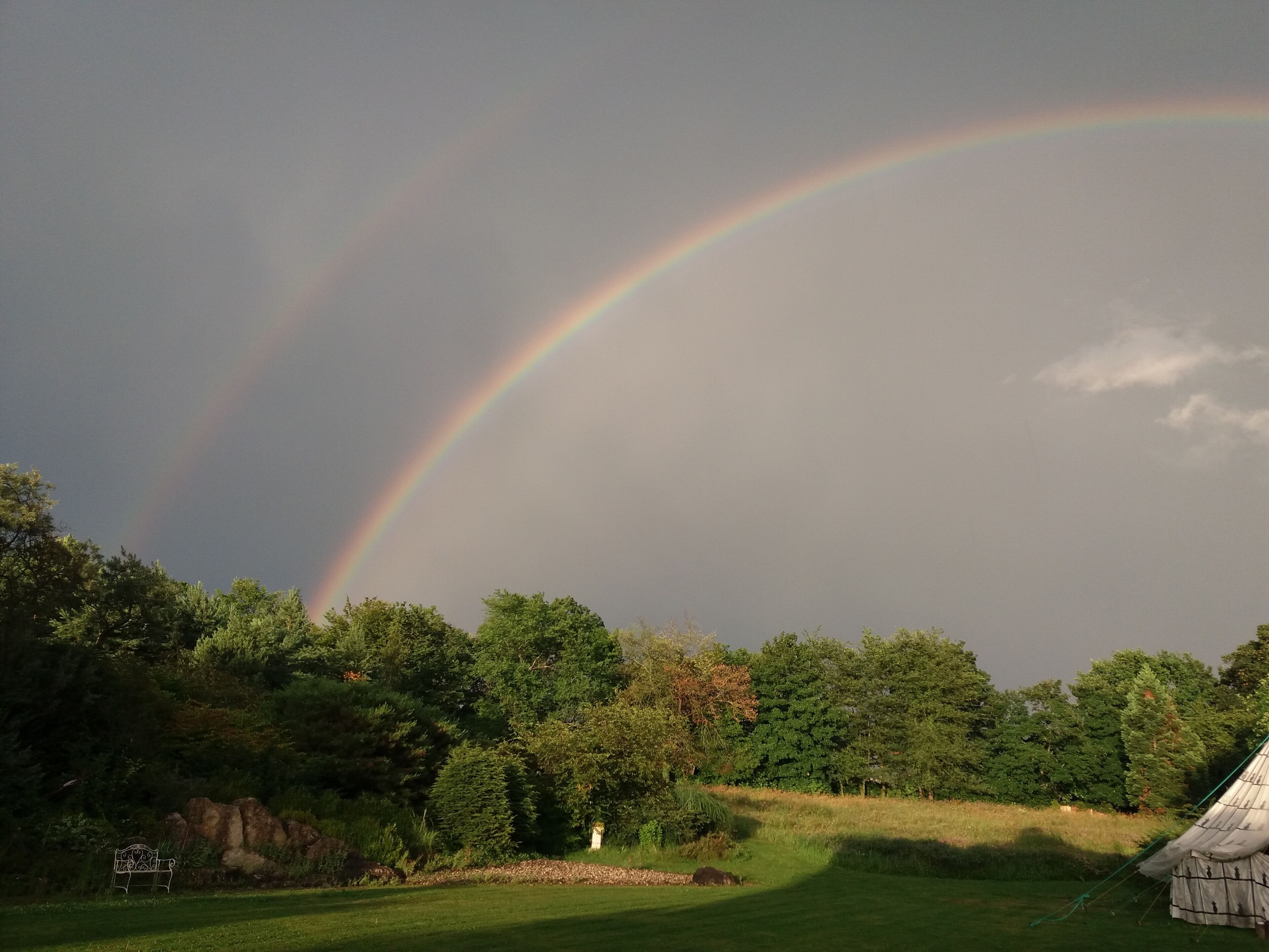 A grassy field with a white metal bench, a stone structure, and a rainbow in the sky.