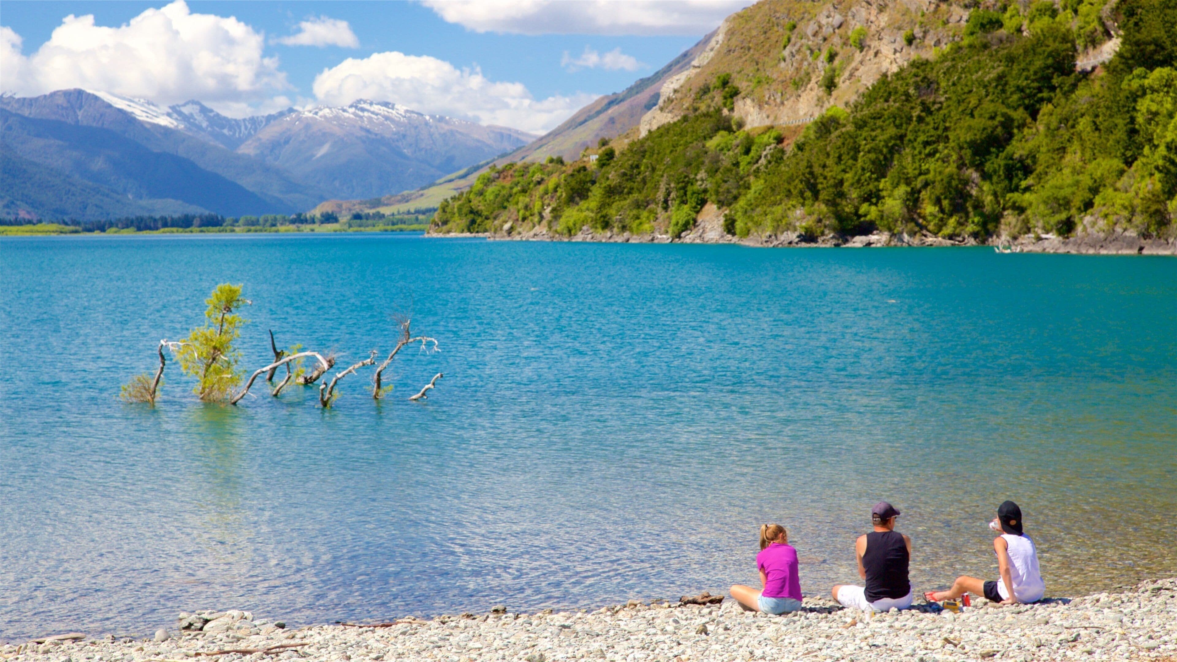 A lake with a rocky shoreline, a few people sitting by the water, and mountains in the background.
