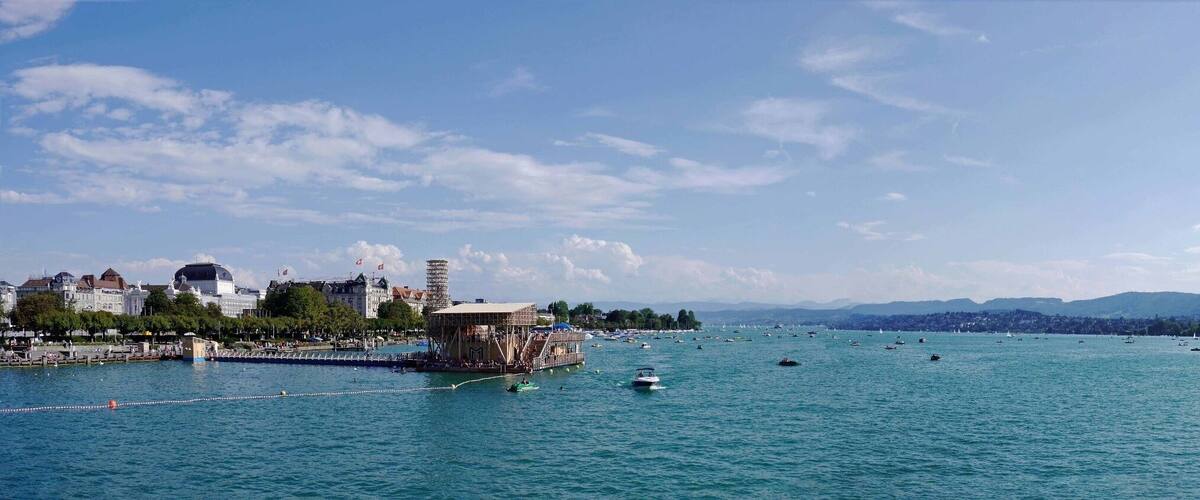 A waterfront cityscape with a prominent tower, boats, and a distant mountain range.