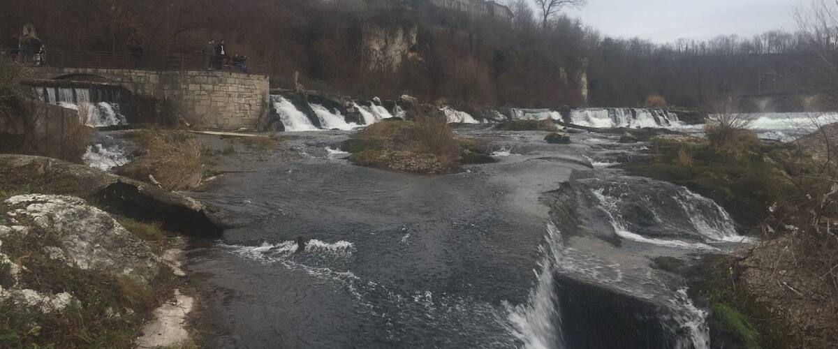 A waterfall cascading over rocks, a stone bridge, and buildings in the background.