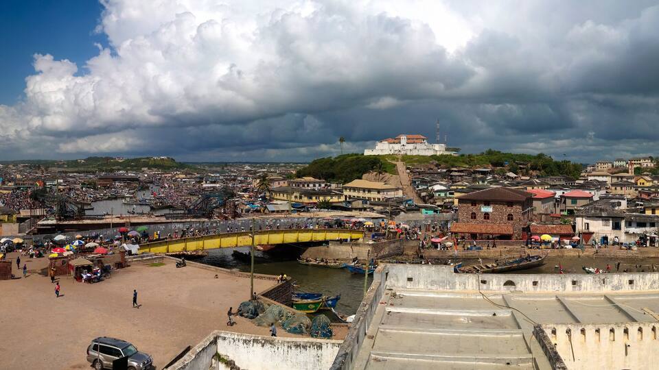 A coastal cityscape with a prominent fort, a bustling market area, and a bridge connecting different parts of the city.