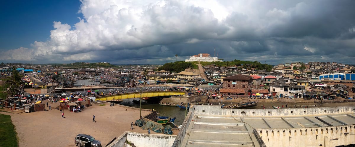 A coastal cityscape with a prominent fort, a bustling market area, and a bridge connecting different parts of the city.