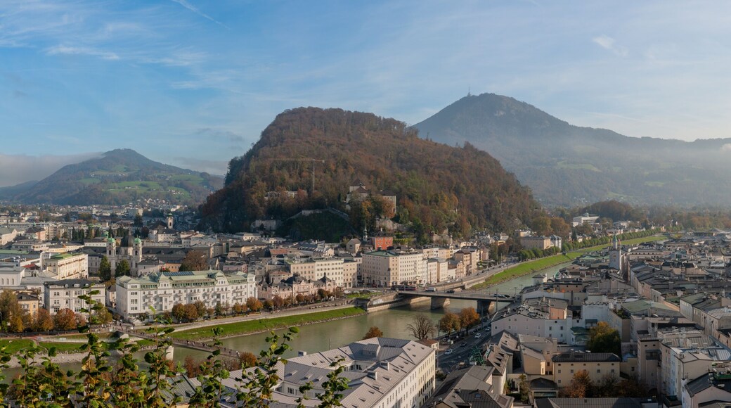 Panoramic view of Salzburg from the Monikapforte, showing the historic old town, the Salzach River, the Mönchsberg hill and Hohensalzburg Fortress under soft morning light.