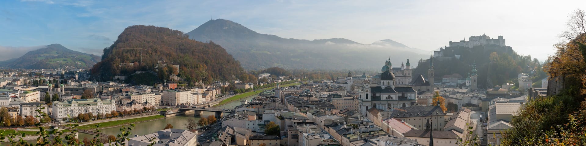 Panoramic view of Salzburg from the Monikapforte, showing the historic old town, the Salzach River, the Mönchsberg hill and Hohensalzburg Fortress under soft morning light.