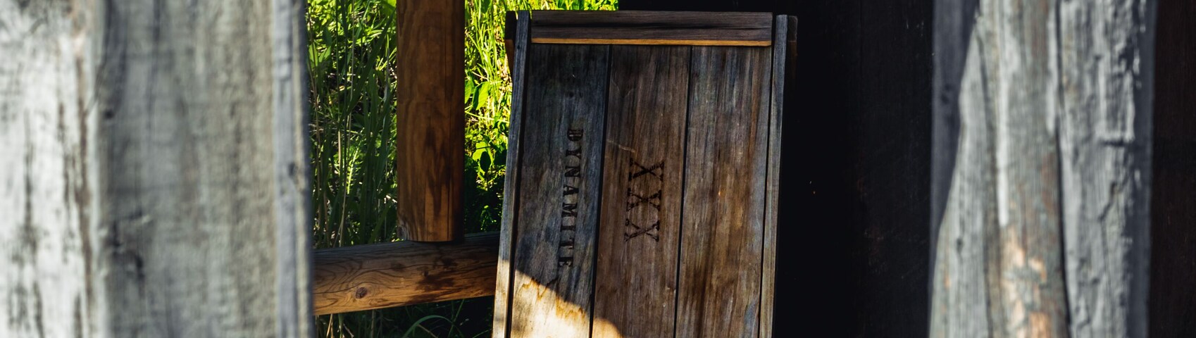 Baú de madeira com 'XXX' e 'PERIGO' em sua superfície, apoiado em um convés de madeira com uma trepadeira verde acima.