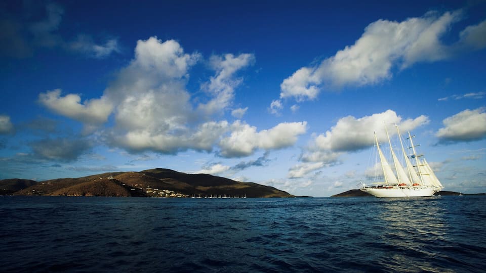 A sailboat on a calm sea with a mountainous coastline and a clear sky.