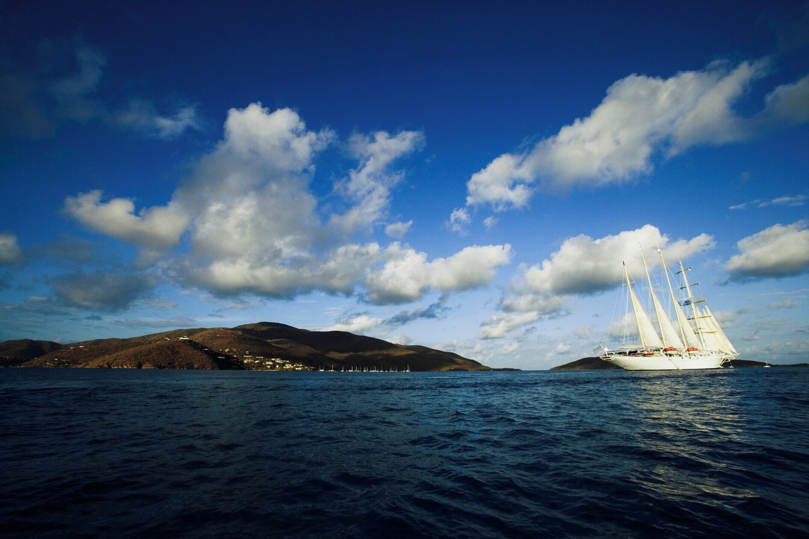 A sailboat on a calm sea with a mountainous coastline and a clear sky.