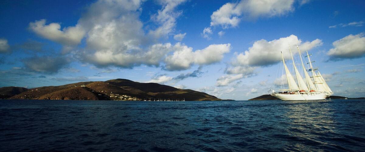 A sailboat on a calm sea with a mountainous coastline and a clear sky.