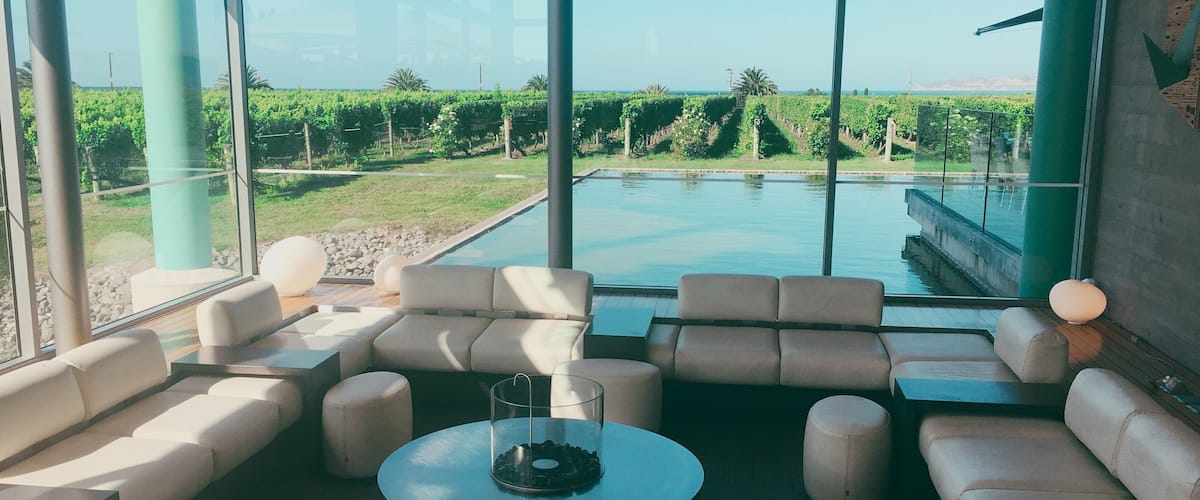 A modern lounge with a pool view, featuring sofas, a coffee table, and a glass wall overlooking a vineyard.