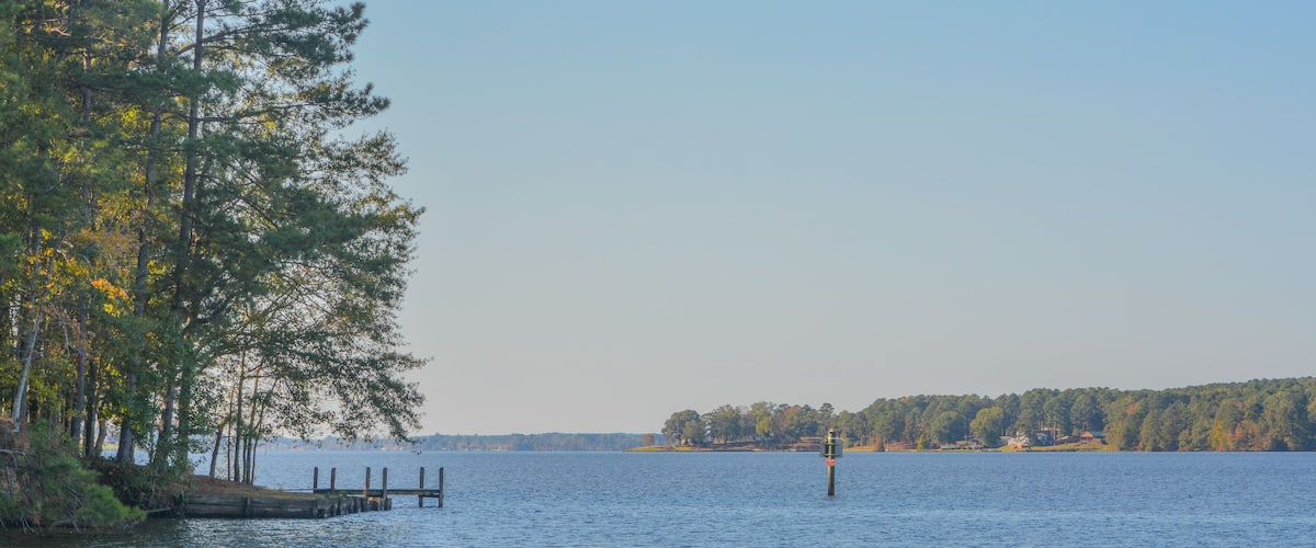 A calm lake with a dock extending into the water, surrounded by trees and a distant shoreline with buildings.