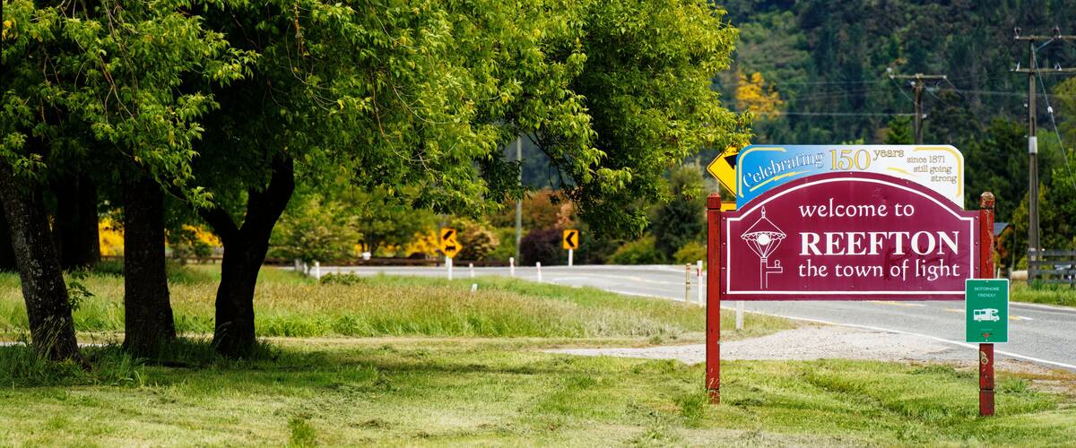A welcome sign for Reefton, a town of light, with a background of lush greenery and a mountainous landscape.