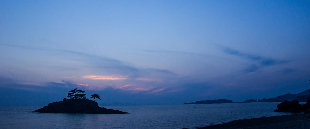 A calm sea at dusk with a small island and distant landforms.